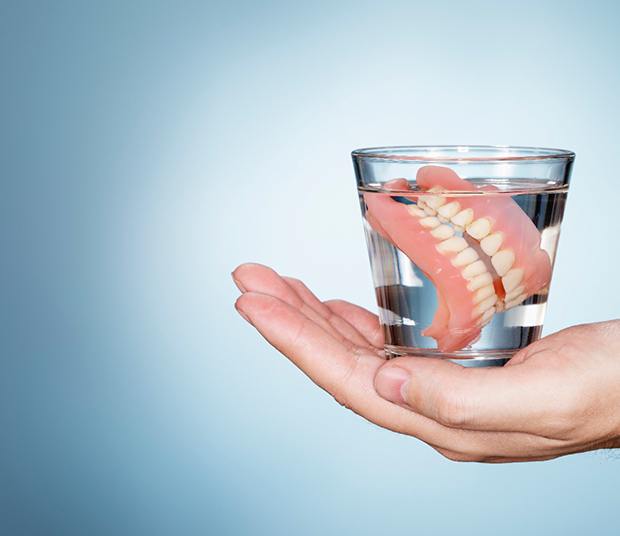 A hand holding a glass of clear fluid and a full set of dentures